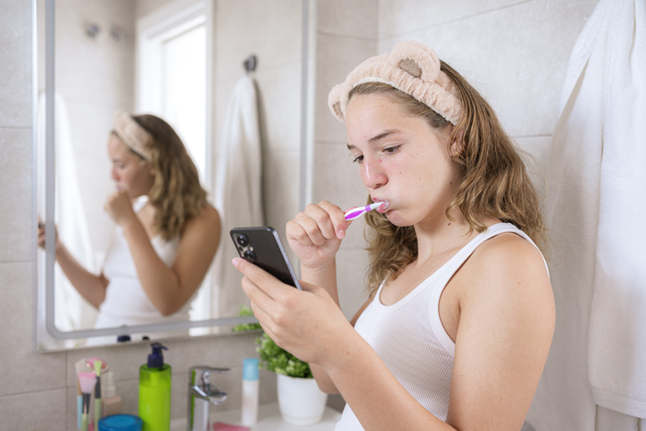 Teenage girl brushing her teeth and using mobile phone in the bathroom. High resolution 42Mp indoors digital capture taken with SONY A7rII and Zeiss Batis 40mm F2.0 CF lens