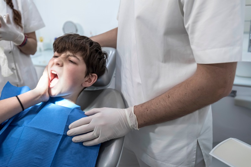 Worried looking boy in the dentist chair in front of the doctor is complaining because of her gum pain and holding her hand on the cheek.